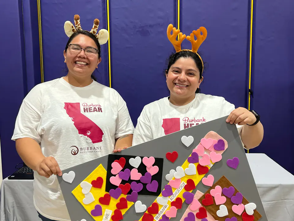 Two members of our team smiling at a community event in playful antler and giraffe headbands, holding a poster covered in paper hearts